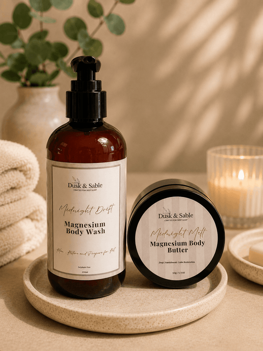 Amber glass pump bottle and jar of Magnesium Body Wash and Body Butter on round wooden tray with white towel and green plant .